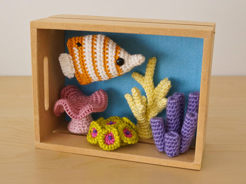 a diorama of crocheted corals and an amigurumi butterflyfish with a blue background inside a wooden crate
